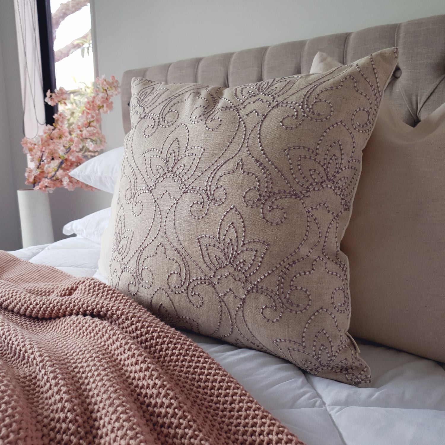 Decorative Threads Indie cushion with floral pattern on a bed with pink blanket and flowers in the background