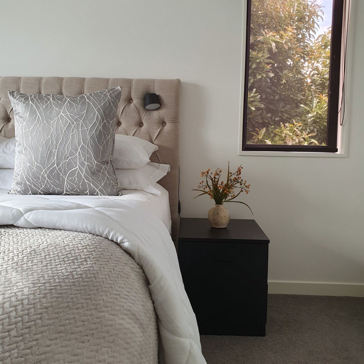 Neatly made bed with grey silver cushion and a vase of flowers on a nightstand next to a window.
