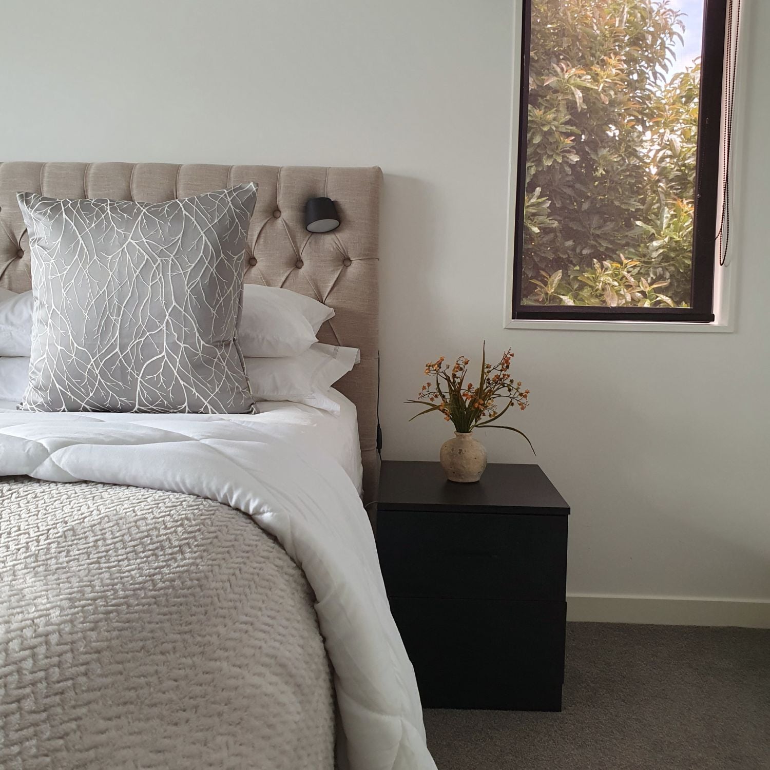 Neatly made bed with grey silver cushion and a vase of flowers on a nightstand next to a window.