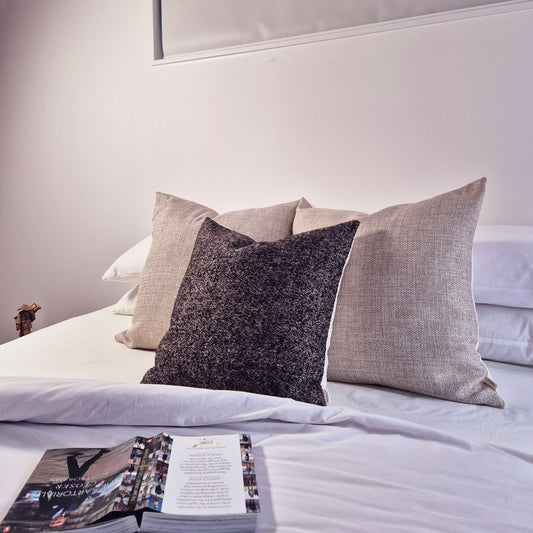 Decorative Threads Herringbone Harmony cushion combo in colour natural/chocolate on a bed with Bed with open magazine on a light-colored background.