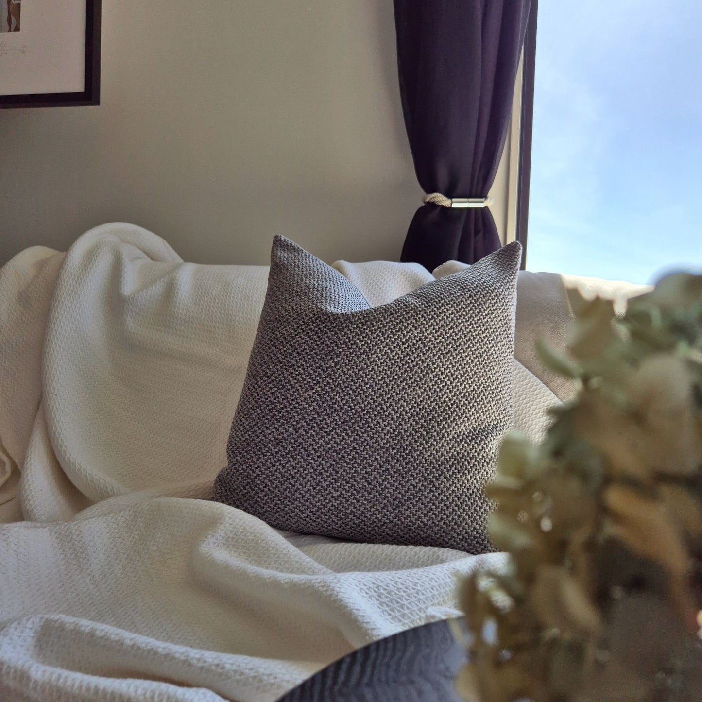 Decorative Threads Larkin cushion in storm grey colour on a couch with a throw and blurred flowers in the foreground.