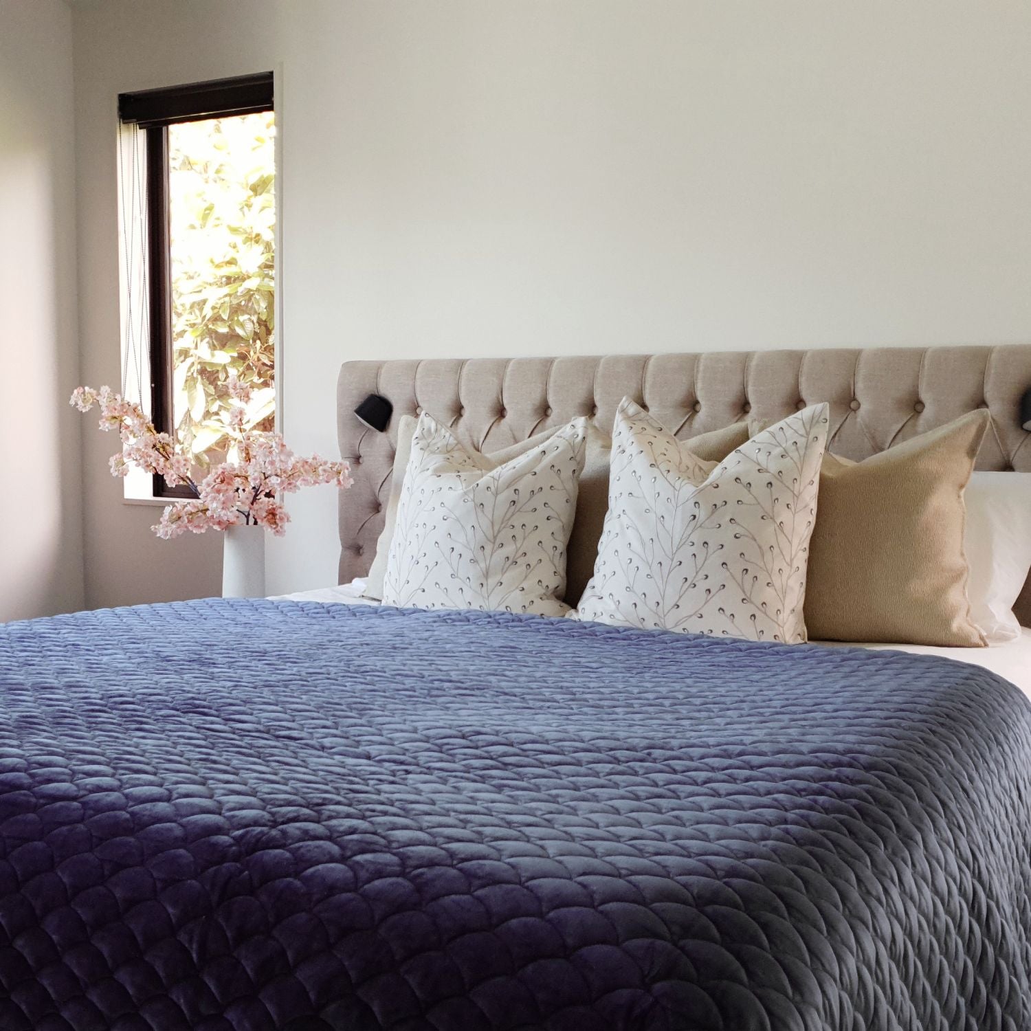 Decorative Threads Twiggy and Katama cushions on a bed featuring a blue quilt, near a window with a plant.